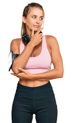 Young blonde woman wearing gym clothes and using headphones with hand on chin thinking about question, pensive expression. smiling with thoughtful face. doubt concept.