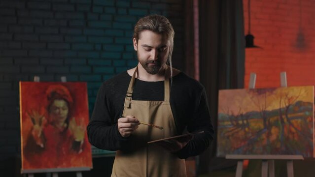 Front view of talented caucasian man in apron mixing with brush paints on wooden color palette. Bearded male painter standing in art studio, smiling and looking at camera.
