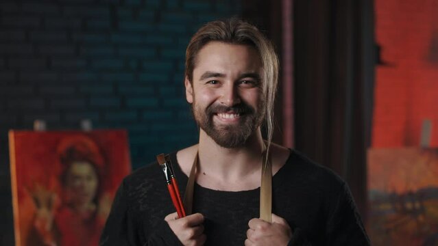 Portrait of handsome male artist posing at modern art studio with sincere smile on face. Caucasian man with trendy beard and haircut holding paintbrushes in hands.