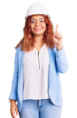 Young latin woman wearing architect hardhat showing and pointing up with finger number one while smiling confident and happy.