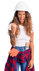 Young hispanic woman with tattoo wearing hardhat and builder clothes angry and mad raising fist frustrated and furious while shouting with anger. rage and aggressive concept.