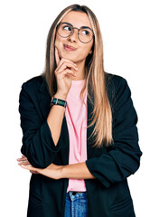 Hispanic young woman wearing business jacket and glasses with hand on chin thinking about question,...