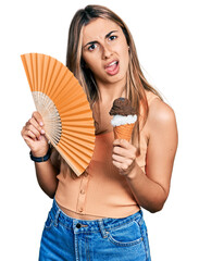 Hispanic young woman holding hand fan eating ice cream in shock face, looking skeptical and sarcastic, surprised with open mouth