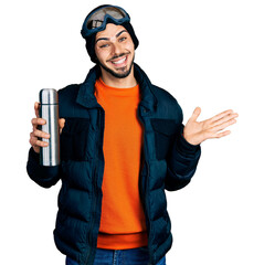 Young hispanic man with beard wearing snow wear and sky glasses holding thermo celebrating achievement with happy smile and winner expression with raised hand