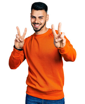 Young hispanic man with beard wearing casual orange sweater smiling looking to the camera showing fingers doing victory sign. number two.