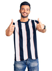 Fototapeta premium Young handsome hispanic man wearing striped tshirt approving doing positive gesture with hand, thumbs up smiling and happy for success. winner gesture.