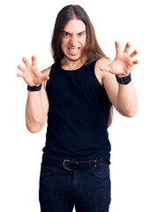 Young adult man with long hair wearing goth style with black clothes smiling funny doing claw...