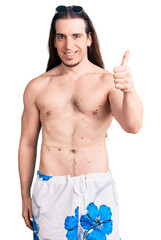 Fototapeta premium Young adult man with long hair wearing swimwear shirtless smiling happy and positive, thumb up doing excellent and approval sign