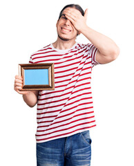 Young adult man with long hair holding empty frame stressed and frustrated with hand on head, surprised and angry face