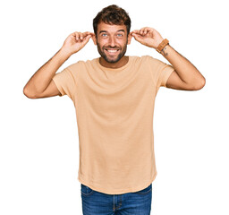 Handsome young man with beard wearing casual tshirt smiling pulling ears with fingers, funny...