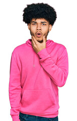 Fototapeta na wymiar Young african american man with afro hair wearing casual pink sweatshirt looking fascinated with disbelief, surprise and amazed expression with hands on chin
