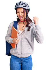 Young beautiful chinese girl wearing student backpack and bike helmet holding book screaming proud, celebrating victory and success very excited with raised arms