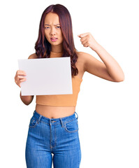 Fototapeta premium Young beautiful chinese girl holding paper banner with blank space annoyed and frustrated shouting with anger, yelling crazy with anger and hand raised