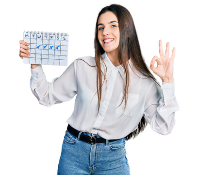 Young brunette teenager holding travel calendar doing ok sign with fingers, smiling friendly gesturing excellent symbol