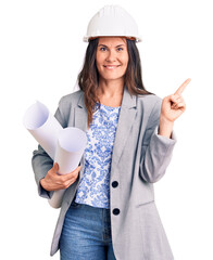 Young beautiful brunette woman wearing architect hardhat holding blueprint smiling happy pointing with hand and finger to the side