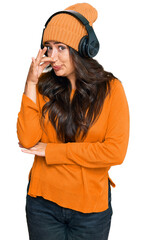 Beautiful brunette young woman listening to music using headphones smelling something stinky and disgusting, intolerable smell, holding breath with fingers on nose. bad smell
