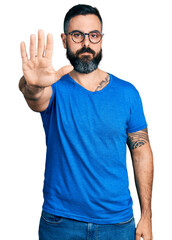 Hispanic man with beard wearing casual t shirt and glasses doing stop sing with palm of the hand. warning expression with negative and serious gesture on the face.