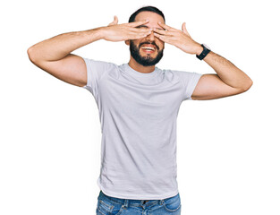 Young man with beard wearing casual white t shirt covering eyes with hands smiling cheerful and funny. blind concept.