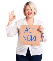 Young blonde plus size woman holding act now cardboard banner doing ok sign with fingers, smiling friendly gesturing excellent symbol