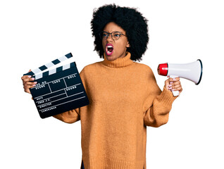 Young african american woman holding video film clapboard and megaphone angry and mad screaming...