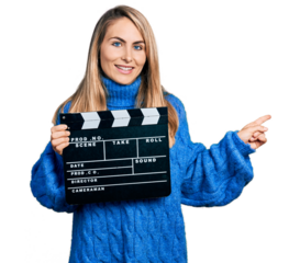 Young blonde woman holding video film clapboard smiling happy pointing with hand and finger to the side