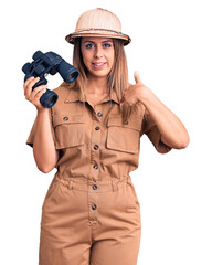 Young beautiful woman wearing explorer hat holding binoculars pointing finger to one self smiling happy and proud