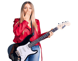 Young beautiful blonde woman playing electric guitar covering mouth with hand, shocked and afraid...