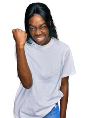Young african american woman wearing casual white t shirt angry and mad raising fist frustrated and furious while shouting with anger. rage and aggressive concept.