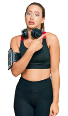 Beautiful blonde woman wearing gym clothes and using headphones pointing aside worried and nervous with forefinger, concerned and surprised expression