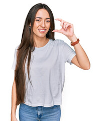 Young hispanic girl wearing casual white t shirt smiling and confident gesturing with hand doing small size sign with fingers looking and the camera. measure concept.