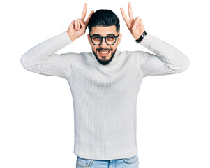 Fototapeta premium Young arab man with beard wearing elegant turtleneck sweater and glasses posing funny and crazy with fingers on head as bunny ears, smiling cheerful