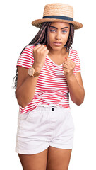 Young african american woman with braids wearing summer hat ready to fight with fist defense gesture, angry and upset face, afraid of problem