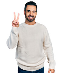 Naklejka premium Young hispanic man with beard wearing casual white sweater showing and pointing up with fingers number two while smiling confident and happy.