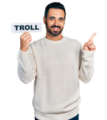 Young hispanic man with beard holding troll banner smiling happy pointing with hand and finger to the side