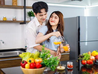 Asian young lovely couple handsome husband standing at kitchen counter full of fresh fruits and vegetables hugging beautiful wife from behind while pouring organic orange juice from jug to glass