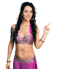 Young indian woman wearing belly dancer costume with a big smile on face, pointing with hand finger to the side looking at the camera.