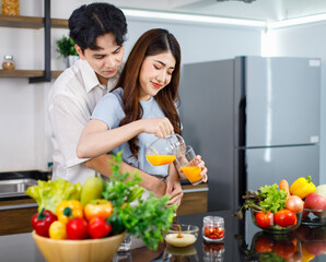 Asian young lovely couple handsome husband standing at kitchen counter full of fresh fruits and vegetables hugging beautiful wife from behind while pouring organic orange juice from jug to glass