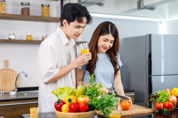 Asian young lovely couple handsome husband standing at kitchen counter full of fresh fruits and vegetables holding glass of orange juice ready to drink while beautiful wife cutting tomato with knife