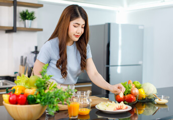 Asian young beautiful housewife standing at kitchen counter full of organic fresh fruits and vegetables bowl using knife preparing cutting red and green apple on chopping board ready to serve on dish