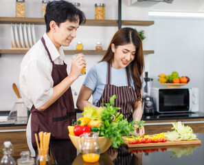 Asian young lovely couple handsome husband standing at kitchen counter full of fresh fruits and vegetables holding glass of orange juice ready to drink while beautiful wife cutting tomato with knife
