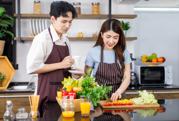 Asian wife in stripe apron stand smiling at kitchen counter full of organic fresh fruits and vegetables in bowl preparing cutting salad ingredients on chopping board while husband drinking hot coffee