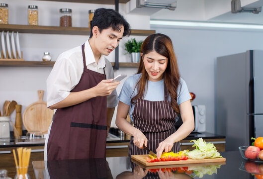 Asian millennial young lover couple handsome husband using smarphone taking photo of pretty wife in apron standing smiling cutting fresh raw organic vegetables on chopping board with knife in kitchen