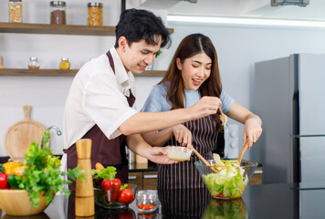 Asian young cheerful beautiful housewife in stripe apron standing smiling at kitchen counter using wood utensils mixing organic fresh salad vegetables in bowl while husband help adding cream dressing