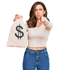 Young brunette woman holding dollars bag pointing with finger to the camera and to you, confident...