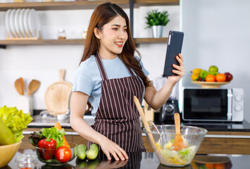 Portrait shot Asian young cheerful housewife in apron wearing play touchscreen tablet computer standing smiling behind kitchen counter