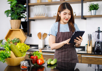 Obraz na płótnie Canvas Portrait shot Asian young cheerful housewife in apron wearing play touchscreen tablet computer standing smiling behind kitchen counter