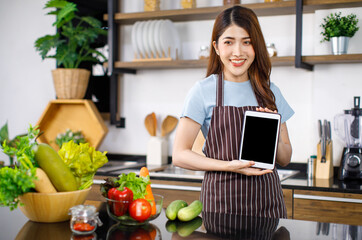 Asian young lovely housewife in apron standing smiling holding presenting showing black blank screen tablet computer with advertising copy space behind kitchen counter with fresh organic vegetables