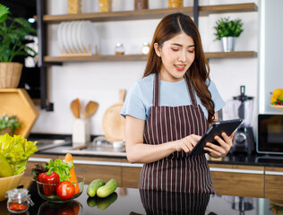 Obraz na płótnie Canvas Portrait shot Asian young cheerful housewife in apron wearing play touchscreen tablet computer standing smiling behind kitchen counter