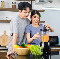 Asian young romantic lover couple handsome husband standing smiling hugging beautiful wife from behind while using blender blending preparing mixed fresh raw organic fruits juice on kitchen counter
