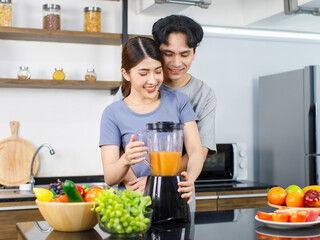 Asian young romantic lover couple handsome husband standing smiling hugging beautiful wife from behind while using blender blending preparing mixed fresh raw organic fruits juice on kitchen counter